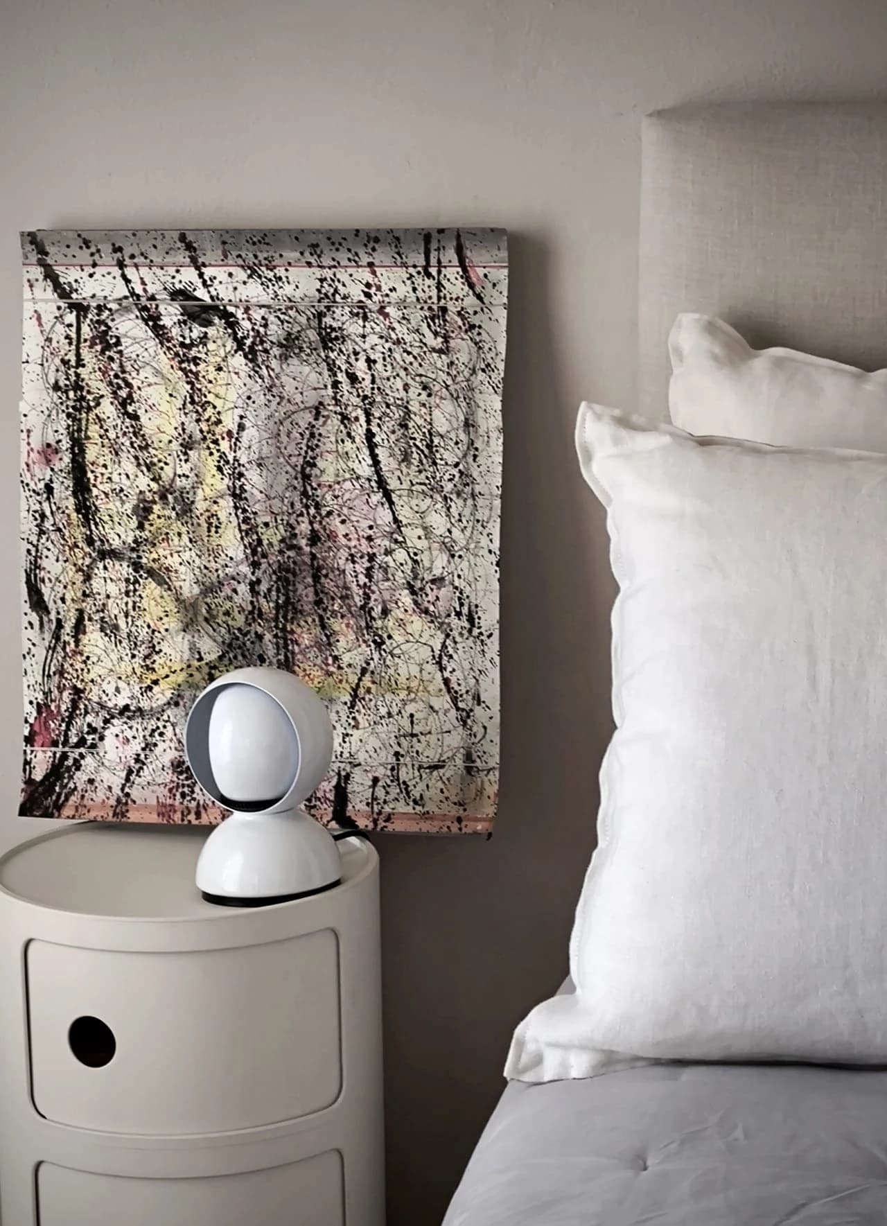 A splattered work on paper above a bedside table and a linen bed